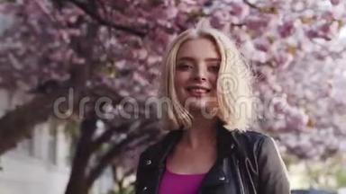 在樱花盛开的城市中漫步的迷人的金发少女的后景，转向镜头微笑